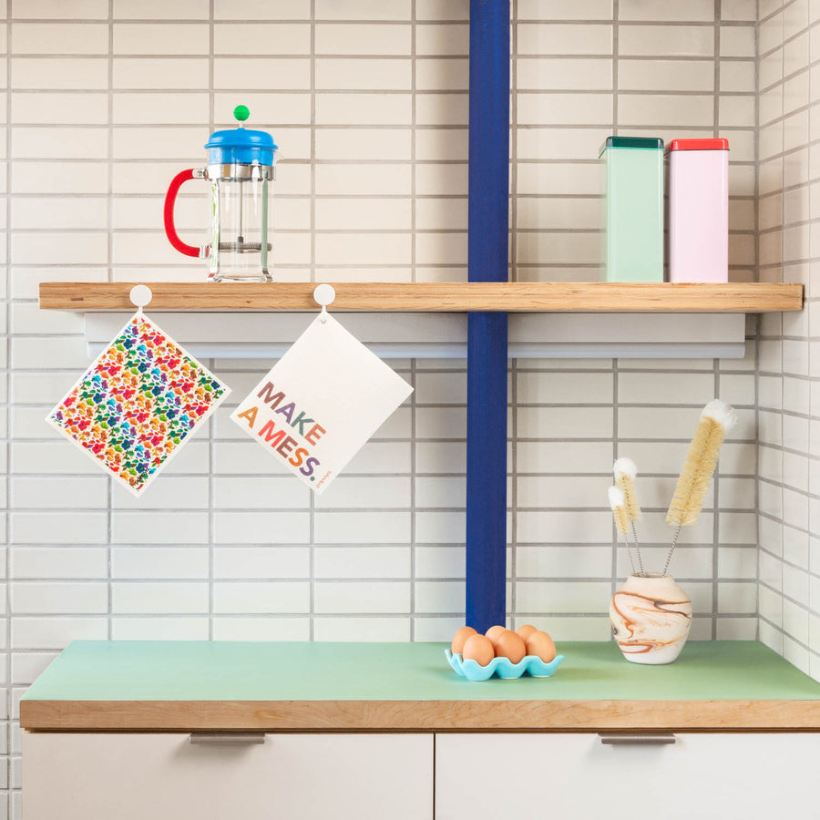Two reusable paper towels hanging on hooks in modern kitchen with colorful floral design and make a mess words 